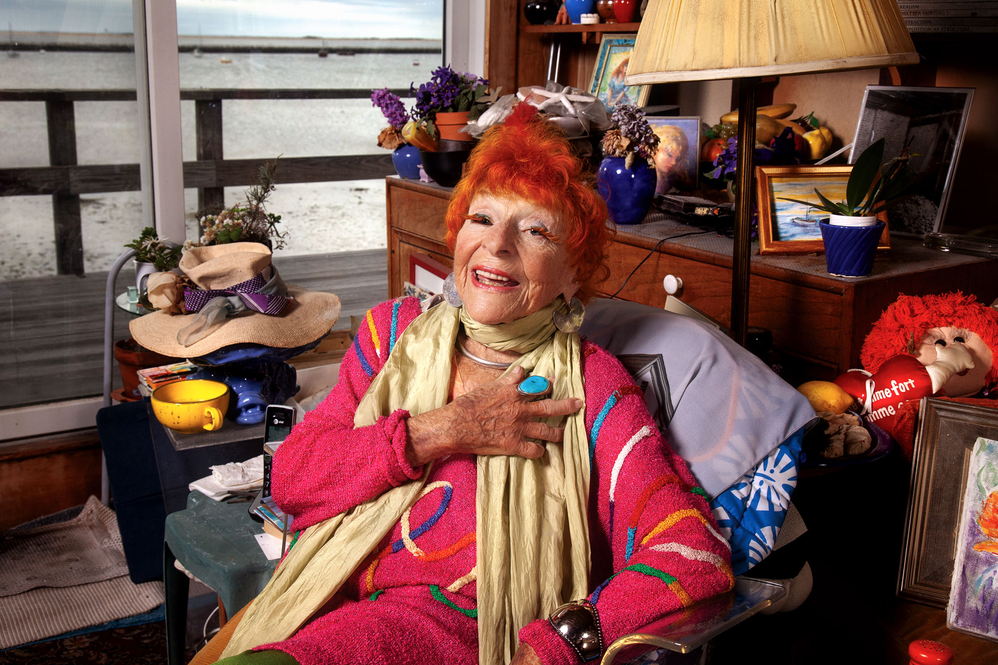 Ilona Royce-Smithkin with bright red hair, clothing photographed at home in Provincetown with a view of the ocean through sliding deck doors behind her.