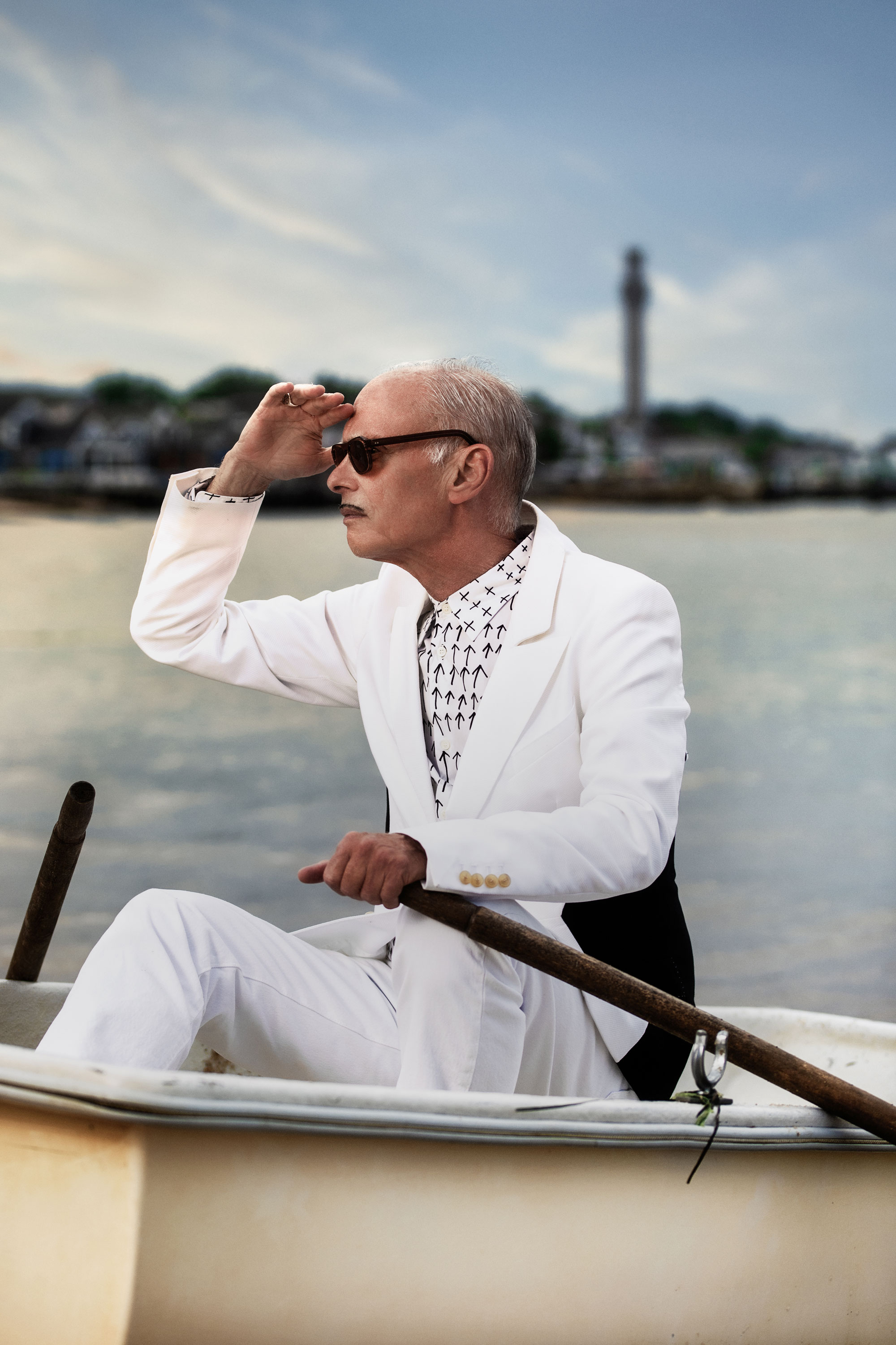 John Waters in a white suit, seated in a dingy on the beach in Provincetown with a the ocean and a lighthouse behind him.