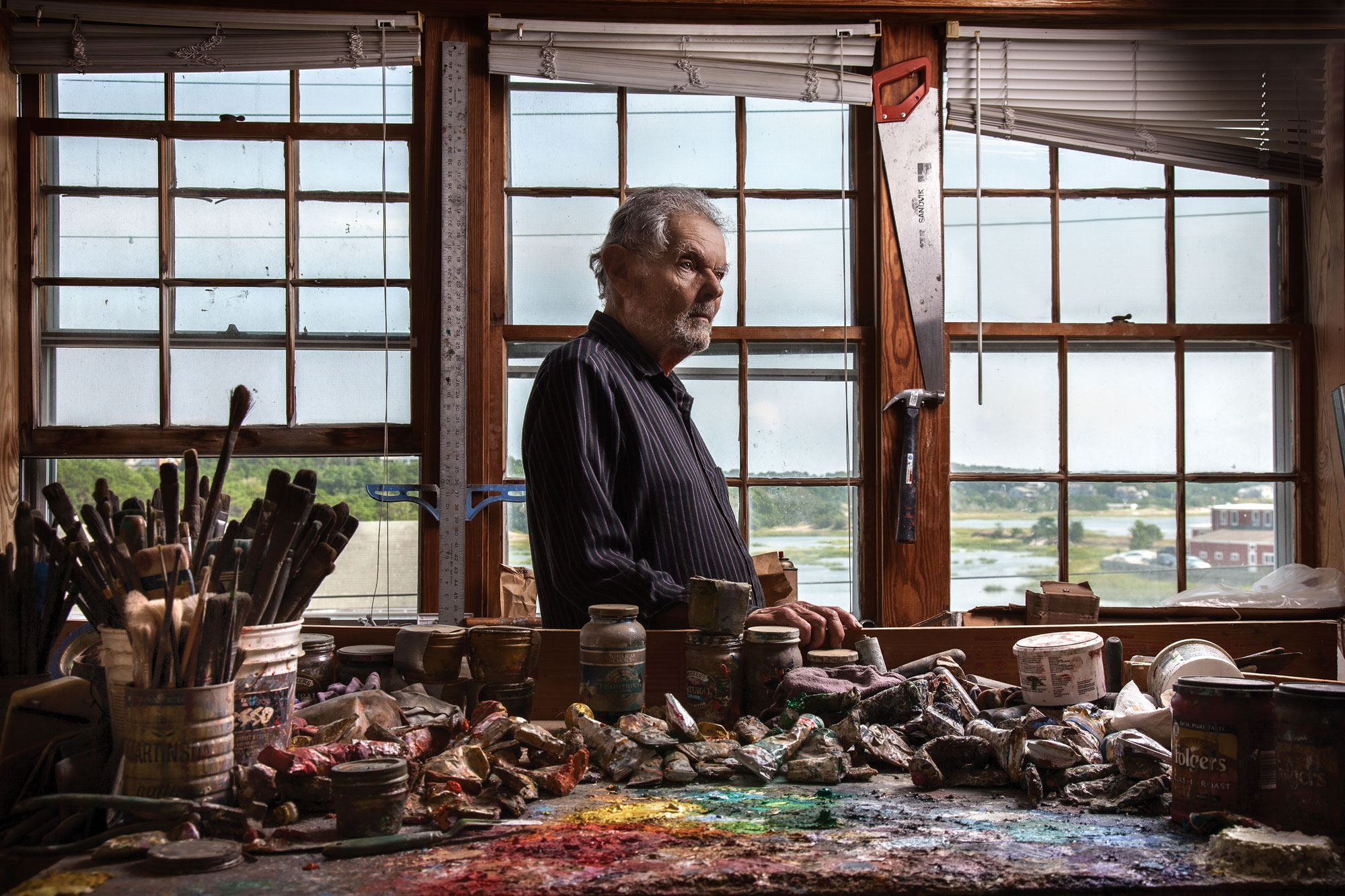 Photo of Robert Henry photographed in his studio behind a table full of paint tubes and brushes with a view of Provincetown through the windows behind him.