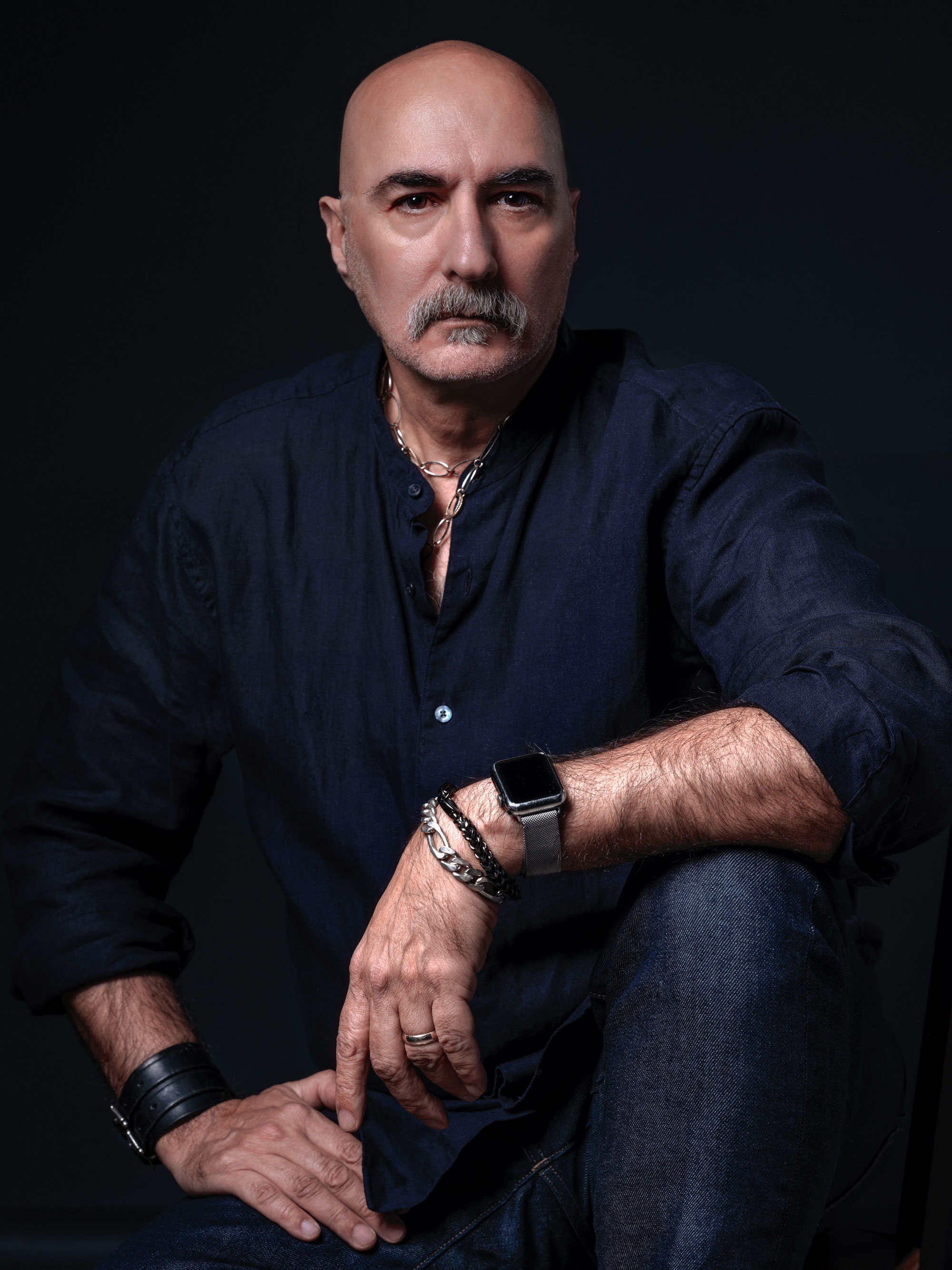 Photo of photographer Ron Amato in a dark blue shirt, dark blue jeans, with silver and leather wristbands and necklaces, seated with a black background